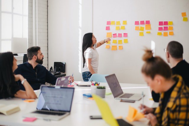 Employees sat at a table with laptops watching a woman explain something on a whiteboard using post-it notes.