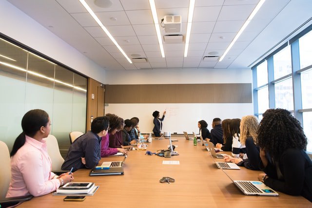 A diverse group of employees at a meeting sat around a table watching somebody stood up explain something on a whiteboard.