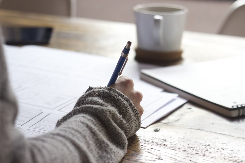 Person writing notes at a table with a warm beverage in front of them